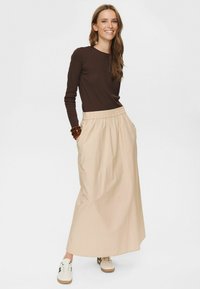 Jupe beige longue jusqu'aux chevilles avec une taille élastique et des poches latérales. Associée à un haut ajusté à manches longues marron et des baskets blanches décontractées.