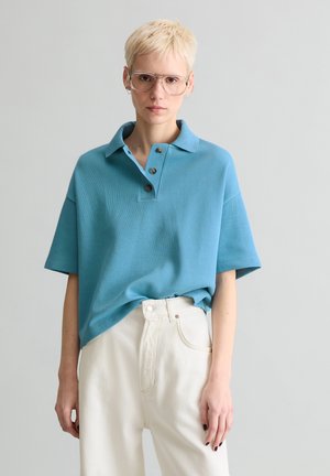 Person mit kurzem blondem Haar, der ein übergroßes blaues Poloshirt, weiße, hoch taillierte Hosen und große, klarsichtige Brillenfassungen vor einem einfarbigen Hintergrund trägt.