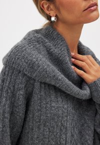 Szary sweter z dzianiny z szerokim, opadającym kołnierzem i prążkowaną teksturą. Ręka modelki spoczywa na kołnierzu, eksponując miękki materiał.