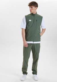 Zelená lehká vesta s vysokým límcem, se zipovými kapsami, spárovaná s odpovídajícími zelenými kalhotami a bílými teniskami. Hladká, prodyšná tkanina.