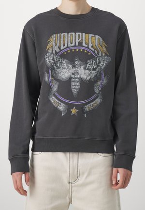 Person trägt dunkelgraues Sweatshirt mit großem Mottenmotiv, Sternen und dem Text "KOOPLESS" in stilisierter Schrift auf der Vorderseite.