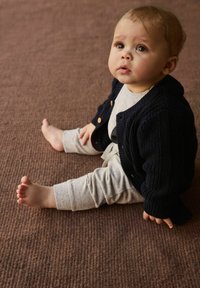 Spædbarn iført en marineblå strikket cardigan, lysegrå skjorte og lysegrå mønstrede bukser, siddende på en tekstureret brun overflade.