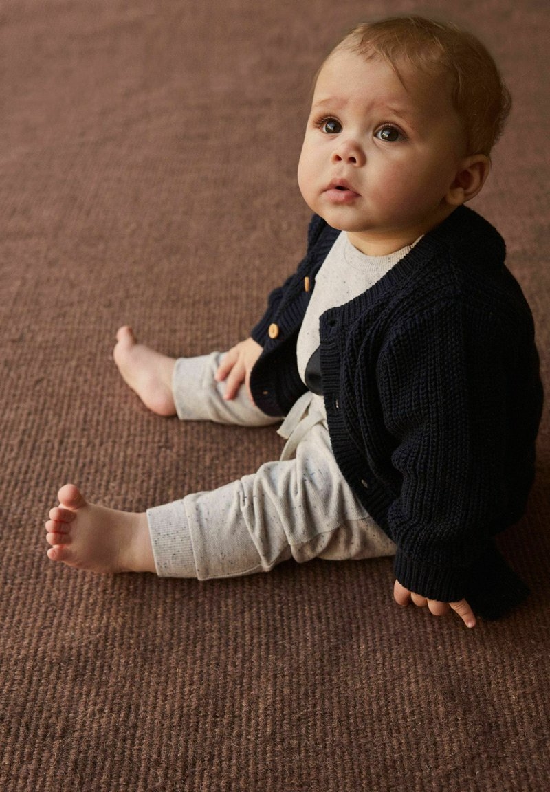 Spædbarn iført en marineblå strikket cardigan, lysegrå skjorte og lysegrå mønstrede bukser, siddende på en tekstureret brun overflade.