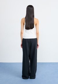 Una mujer con cabello largo lleva una blusa sin mangas blanca y pantalones negros sueltos, de espaldas contra un fondo claro y una alfombra azul.