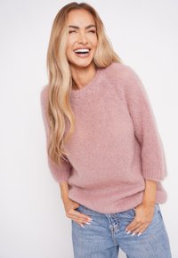 Uśmiechaj�ąca się kobieta z długimi blond włosami, ubrana w różowy puszysty sweter i niebieskie dżinsy, stojąca na tle jednolitej białej ściany.