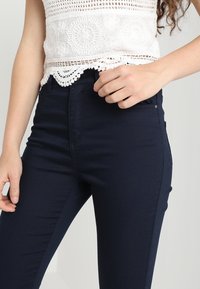 Vrouw in donkerblauwe, strakke broek en een witte gehaakte crop top, met één hand bij de taille.