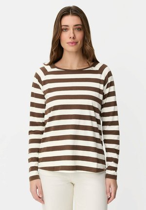 Kobieta o brązowych włosach, ubrana w brązowo-biały, poziomo paskowany, długi rękaw oraz kremowe spodnie, stojąca na jednolitym tle.