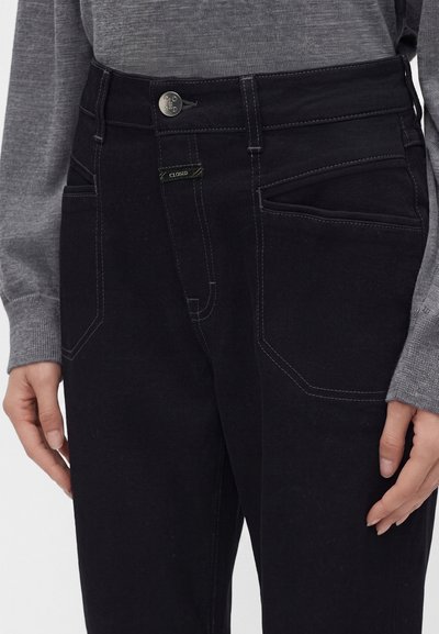 Gros plan d’un jean noir taille haute avec coutures visibles, un bouton en métal, petite étiquette logo, porté avec un haut gris à manches longues.