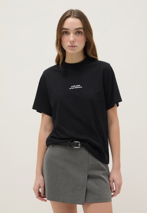 Schwarzes Baumwoll-T-Shirt mit kurzen Ärmeln, bedruckt mit weißem Text "pretty good at bad decisions" auf der Brust, kombiniert mit einem grauen Rock.