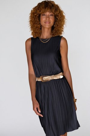 Vrouw met krullend haar die een mouwloze zwarte geplooide jurk draagt, textuurt beige riem, gouden ketting en armband, staand tegen een effen achtergrond.