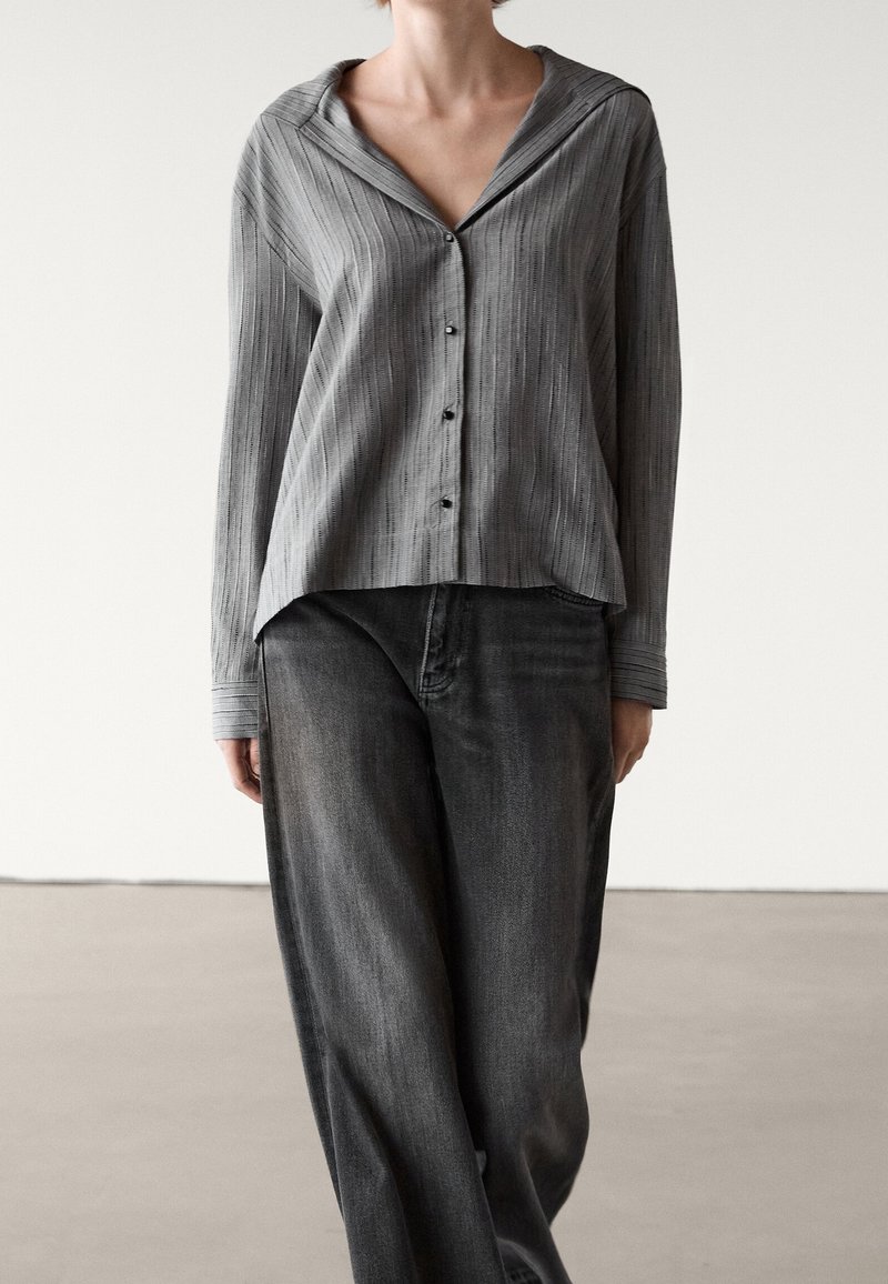 Persona che indossa una camicia grigia morbida con sottili righe e jeans neri a gamba larga, in piedi su uno sfondo chiaro e semplice.