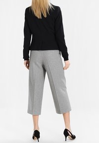 Kvinna iklädd svart långärmad jacka, grå vida cropped byxor och svarta högklackade skor, stående mot vit bakgrund.