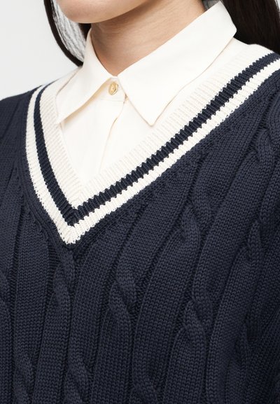 Femme portant un pull à col en V en maille torsadée bleu marine avec des rayures crème et noires, par-dessus une chemise blanche à col.