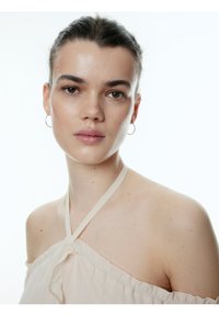 Beige off-shoulder top met een gathered halslijn en een gestrikt detail. Huidtint is licht, met minimalistische make-up en kleine oorringen.