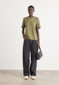 Olivgrüne Baumwoll-T-Shirt mit kurzen Ärmeln, kombiniert mit schwarzen hochgeschnittenen, weiten Jeans. Mit einer kleinen schwarzen Handtasche accessoriert.
