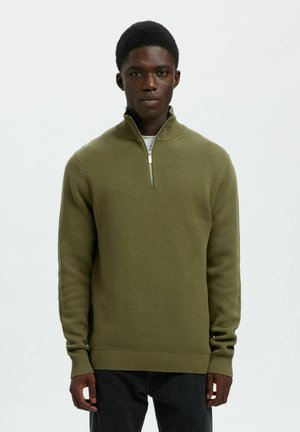 Młody mężczyzna w oliwkowej zielonej bluzie z półzamkiem, noszący białą koszulę i czarne spodnie, stojący na tle białego tła.