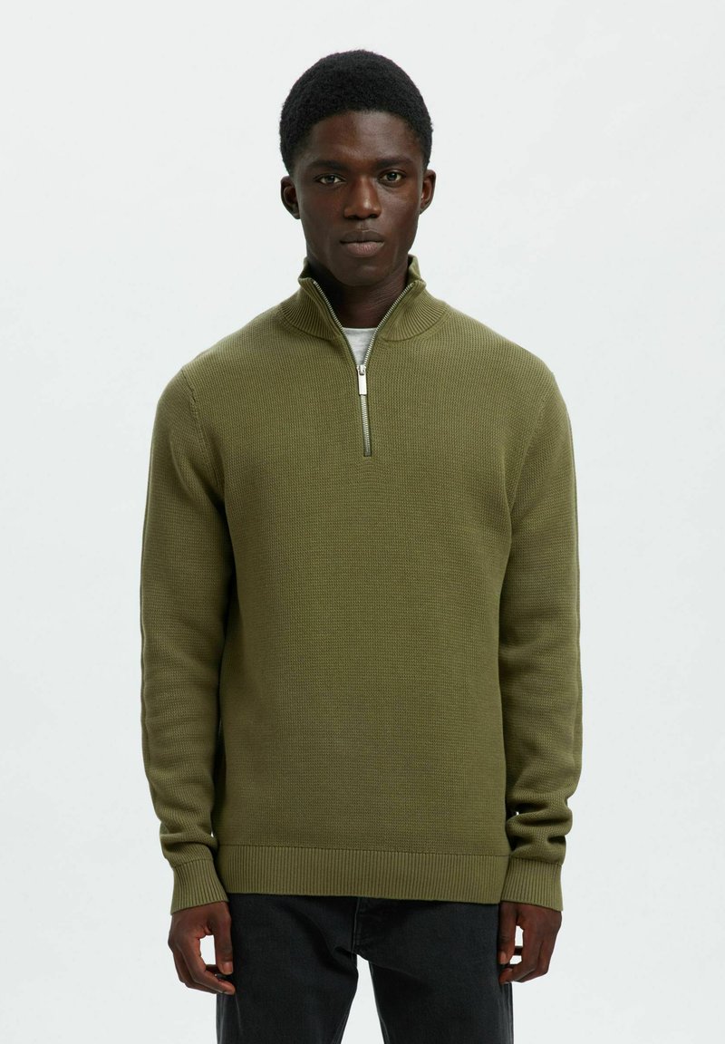 Jaunas vyras dėvi alyvuogių žaliais pusiau užtrauktukais megztinį ant baltos marškinės, juodomis kelnėmis, stovintis prieš paprastą baltą foną.