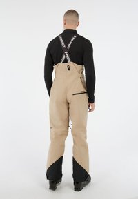 Beige waterdichte sneeuwbroek met verstelbare banden, een gladde textuur en zwarte accenten aan de onderkant. Voorzien van een ritszak aan de achterkant.