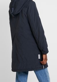 Voděodolná parka v námořnické modři s kapucí, dlouhými rukávy a nastavitelnými manžetami. Má zadní štítek a boční zipy pro ventilaci.