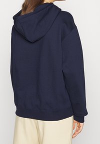 Mörkblå hoodie med avslappnad passform, ribbade muddarna och nederkant, tillverkad av mjukt tyg. Har en känguruficka och sömdekor.