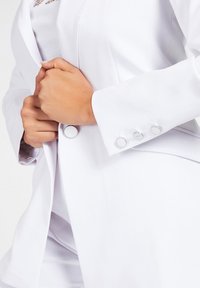 Valkoinen räätälöity bleiseri sileästä kankaasta. Varusteena pyöreät hopeiset napit hihoissa ja edessä, jotka luovat siistin ja jämäkän muotoilun.