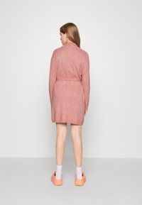 Πλεκτό ροζ μπουφάν με ζώνη, μακριά μανίκια και ραμμένο τελείωμα. Μοντέλο που φοράει ανοιχτόχρωμες κάλτσες και πορτοκαλί παπούτσια, σε θέα από πίσω.