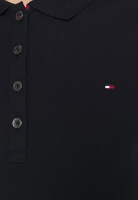Gros plan sur un polo noir avec quatre boutons noirs et un petit logo rectangulaire rouge, blanc et bleu sur la poitrine gauche.