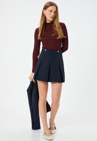 Burgunderfarbener, gerippter Longsleeve-Oberteil kombiniert mit einem marineblauen, plissierten Minirock mit zwei vorderen Knöpfen. Das Model hält eine dunkle Jacke. Helle Schuhe.