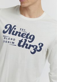 Hvid langærmet skjorte med mørkeblå tekst, der står "EST. Ninetg thrz3 BLEND DENIM." Blødt, afslappet bomuldsstof.