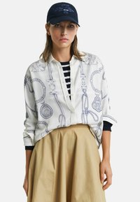 Weißes Blusenshirt mit blauem maritimem Seil-Muster, kombiniert mit einer marineblauen Kappe mit dem Text "New Haven" und einem beigefarbenen, ausgestellten Rock.