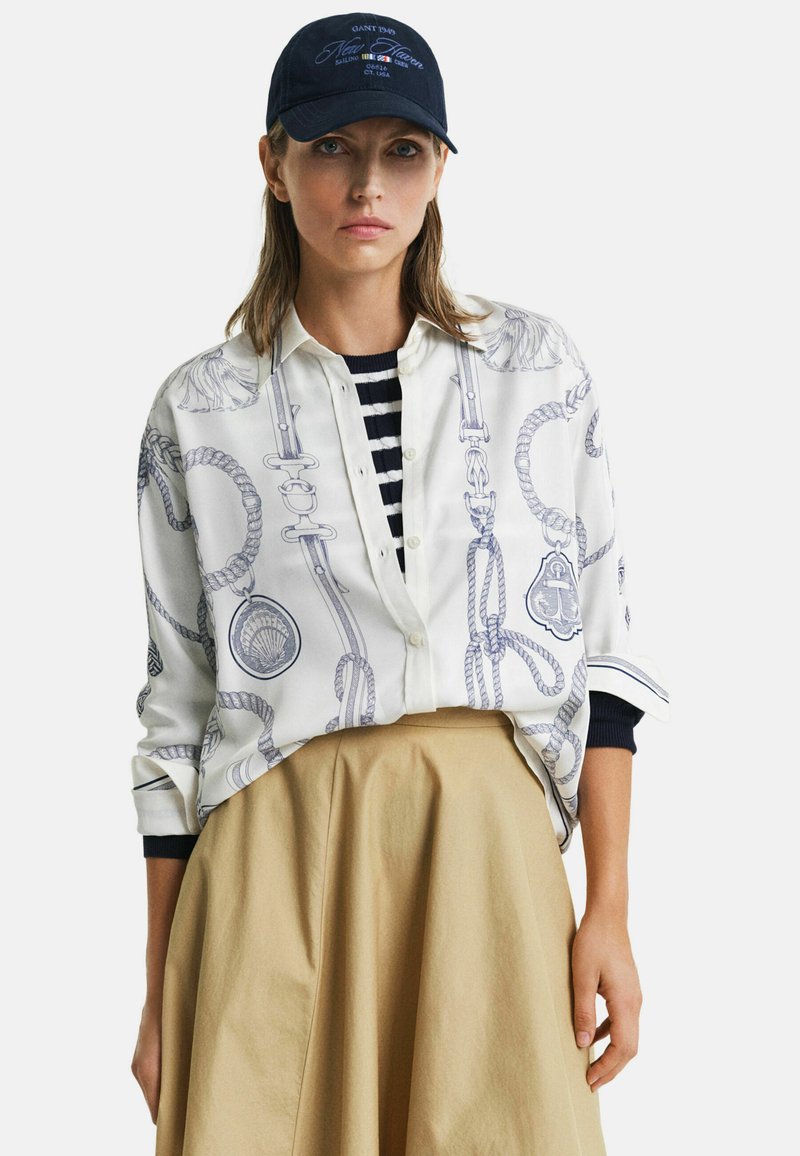 Weißes Blusenshirt mit blauem maritimem Seil-Muster, kombiniert mit einer marineblauen Kappe mit dem Text "New Haven" und einem beigefarbenen, ausgestellten Rock.
