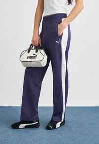 Μωβ φόρμες με λευκές ρίγες στο πλάι, ελαστική ζώνη μέσης και τσέπες, σε συνδυασμό με μαύρα και λευκά αθλητικά Puma και μια μικρή λευκή τσάντα χειρός.