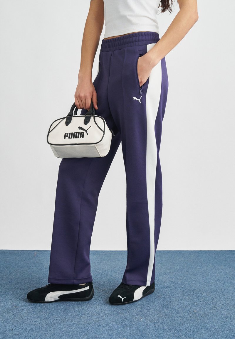 Μωβ φόρμες με λευκές ρίγες στο πλάι, ελαστική ζώνη μέσης και τσέπες, σε συνδυασμό με μαύρα και λευκά αθλητικά Puma και μια μικρή λευκή τσάντα χειρός.