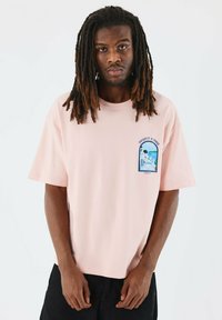 Camiseta de manga corta rosa en tejido suave, con un diseño gráfico en azul y blanco en la parte izquierda del pecho. Ajuste relajado.