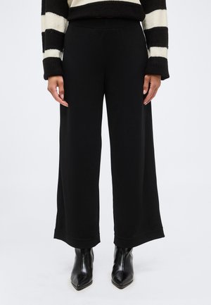 Osoba oblečená v čiernych nohaviciach s širokými nohavicami, čiernych členkových topánkach so špicatou špičkou a čiernobielom pruhovanom pulóvri s dlhými rukávmi.