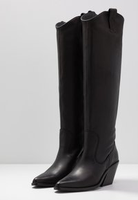 Czarne skórzane kozaki za kolano z ostrym noskiem i koturnem. Charakteryzują się bocznymi pętelkami do ściągania oraz gładką fakturą ze szwami dekoracyjnymi.