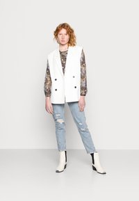 Blazer branco sem mangas com botões duplos, blusa com padrão, jeans desgastados de tom azul claro e botas de tornozelo em creme com detalhes pretos.