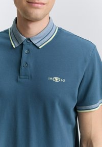 Dunkelblaues Poloshirt mit einem grau gestreiften Kragen, ausgestattet mit einem hellgrünen Akzent und einem gelben Logo auf der Brust. Kurzärmlig, aus Baumwollmaterial.