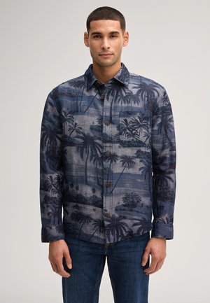 Mężczyzna w długim rękawie, koszuli zapinanej na guziki z niebiesko-czarnym tropikalnym wzorem palm oraz ciemnoniebieskich jeansach, stojący na jednolitym tle.