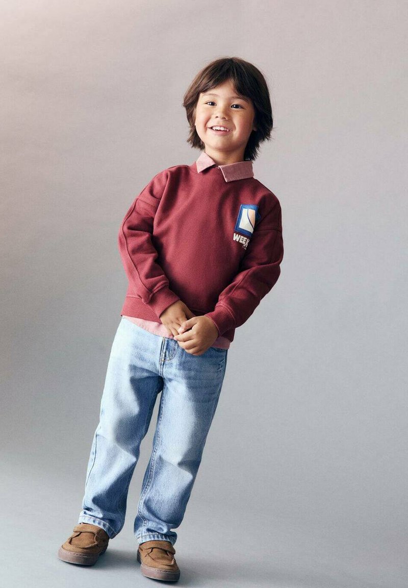Felpa bordeaux con logo, sovrapposta a una camicia rosa con colletto, abbinata a jeans azzurri chiari e scarpe mocassino marroni.