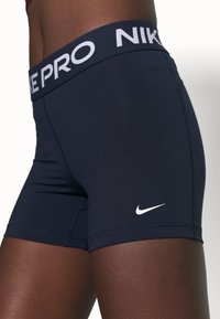 Nike Pro shorts tummansinissä, valmistettu joustavasta kankaasta, jossa on valkoinen logo lahkeessa ja leveä joustava vyötärönauha, jossa on "NIKE PRO" -kirjailu.