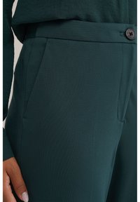 Temno zelene hlače z gladko teksturo, krojene prileganje, stranskimi žepi in enim gumbom za zapenjanje v pasu.