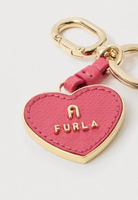 Ροζ δερμάτινο μπρελόκ σε σχήμα καρδιάς με χρυσό περίγραμμα και μεταλλικά μέρη, στερεωμένο σε χρυσό κρίκο κλειδιών, με το λογότυπο Furla σε χρυσά γράμματα.
