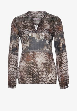 Femme portant un chemisier à manches longues à motifs, un jean noir délavé, des bottes noires, tenant une petite pochette noire.