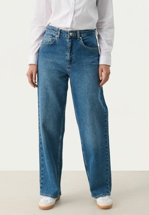 Jean large bleu en tissu denim, avec une taille haute, cinq poches et une texture légèrement décolorée. Associé à des baskets blanches.
