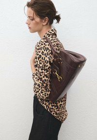 Žena s vlasy sepnutými do drdolu má na sobě leopardí vzorovaný top a černé kalhoty, na rameni nese velkou tmavě hnědou koženou kabelku.