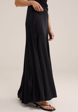 Femeie purtând o fustă lungă și fluidă de culoare neagră, cu talie ajustată, asortată cu sandale negre cu toc mic, stând în fața unui fundal simplu.