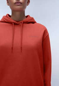 Hoodie in Rostfarbe aus weichem Material, ausgestattet mit einer Kängurutasche, Zugbändern und einem dezenten Logo auf der Brust.