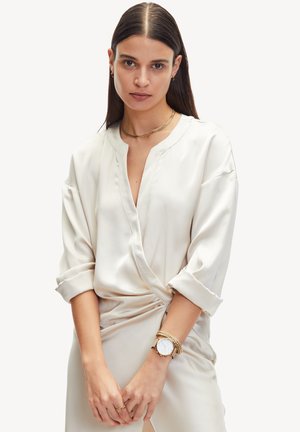 Crème zijden blouse met een diepe V-hals, opgerolde mouwen en een relaxte pasvorm. Gemaakt met gouden accessoires en een horloge. Gladde textuur, elegante draping.