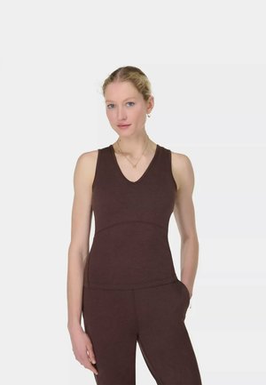 Vrouw met blond haar, gekleed in een bruine mouwloze top en bijpassende broek, staand met één hand in haar zak tegen een eenvoudige lichte achtergrond.